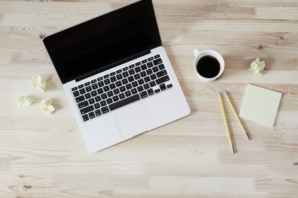 Vue plongeante d'un bureau sur lequel sont posés un ordinateur portable, une tasse à café, un bloc note, un crayon et des feuilles froissées.