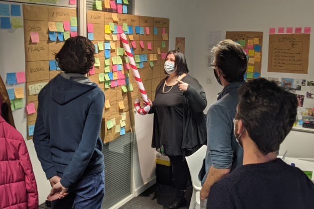 une femme masquée montre des post-it à l'aide d'un sucre d'orge gonflable devant trois développeurs médusés